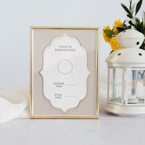 May include: A gold-framed picture with a white background and gold lettering. The text reads "Today is Ramadan Day". There are two blank lines for writing in the times for Suhoor and Iftar.