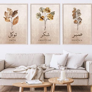Pode incluir: Três impressões de arte botânica emolduradas com fundos bege. Cada impressão apresenta uma ilustração em aquarela de folhas e caules em tons de marrom e dourado. As impressões têm texto árabe e traduções em inglês: Trust, Gratitude e Patience.