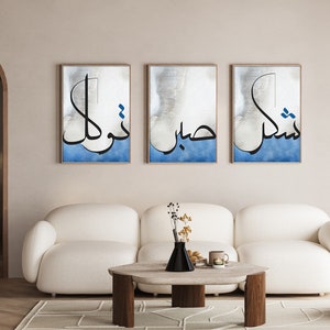 K&ouml;nnte beinhalten: Drei gerahmte Drucke mit arabischer Kalligraphie in Schwarz auf blauem und wei&szlig;em Hintergrund. Die Drucke h&auml;ngen an einer hellbeigen Wand &uuml;ber einem wei&szlig;en Sofa.