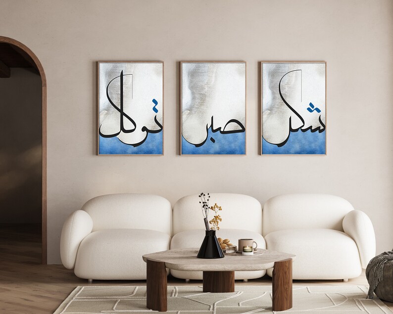 K&ouml;nnte beinhalten: Drei gerahmte Drucke mit arabischer Kalligraphie in Schwarz auf blauem und wei&szlig;em Hintergrund. Die Drucke h&auml;ngen an einer hellbeigen Wand &uuml;ber einem wei&szlig;en Sofa.