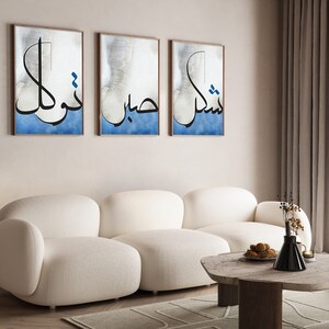K&ouml;nnte beinhalten: Drei gerahmte Drucke mit arabischer Kalligraphie in Schwarz auf blauem Hintergrund. Die Drucke h&auml;ngen an einer hellbeigen Wand &uuml;ber einem wei&szlig;en Sofa. Das Sofa befindet sich in einem Wohnzimmer mit einem Holz-Couchtisch und einer Vase mit Trockenblumen.