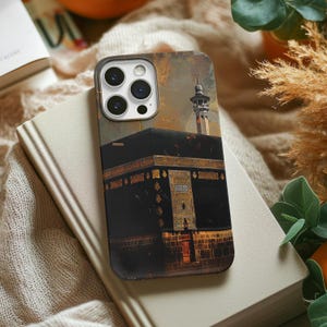 May include: A phone case with an artistic depiction of the Kaaba in Mecca. The case has a black and gold design with a minaret in the background. The case is resting on a book and a beige surface.