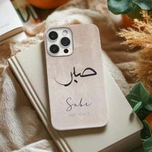 Könnte beinhalten: Eine beige Handyhülle mit einem weißen und schwarzen Design. Die Hülle zeigt das arabische Wort "Sabr" in Kalligraphie, das im Deutschen mit "Geduld" übersetzt wird. Die Hülle hat auch einen Ausschnitt für das Kameraobjektiv und einen schwarzen Rahmen um das Wort "Sabr".
