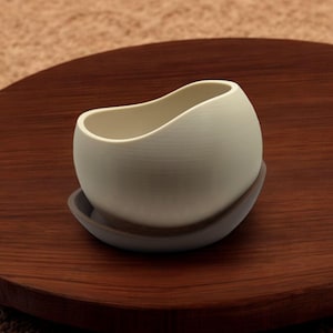 May include: A white ceramic planter with a wavy rim sits on a dark brown wooden coaster. The planter is designed with a rounded, organic shape.