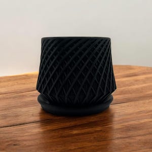 May include: A black geometric planter with a textured, woven design. The planter has a wide base and a narrow top, giving it a unique and modern look.