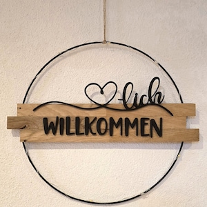 Könnte beinhalten: Ein Holzschild mit dem deutschen Wort „Willkommen“ in schwarzer Schrift. Das Schild ist von einem schwarzen Metallring mit Lichterkette und einer Juteschnur zum Aufhängen umgeben.