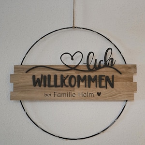 Könnte beinhalten: Ein Holz-Willkommensschild mit dem Wort "Willkommen" in Schwarz, vor einem hellen Holzgrund. Das Schild ist in einem schwarzen Metallring aufgehängt und enthält den Satz "bei Familie Helm" darunter.