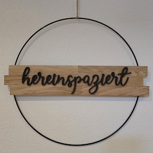 Könnte beinhalten: Ein dekoratives Wandbild mit dem deutschen Wort "hereinspaziert" in Schreibschrift. Der Text ist auf einem Holzbrett montiert, in einem schwarzen Metallring aufgehängt und mit einer natürlichen Schnur befestigt.