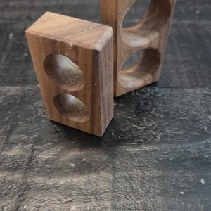 May include: Two rectangular blocks made of brown wood, each featuring two circular cutouts positioned vertically. The wood grain is visible on the surfaces of both blocks, which are oriented upright and stand independently on their bottom edges.