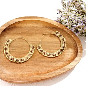 May include: A pair of gold hoop earrings with a beaded design. The earrings are on a wooden surface.