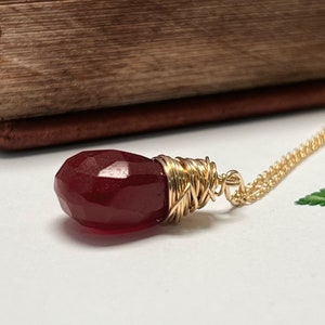 Könnte beinhalten: Eine goldene Kettenkette mit einem roten Edelstein-Anhänger, der in Golddraht gewickelt ist. Der Anhänger ist facettiert und hat eine Tropfenform.