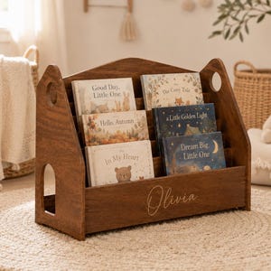 Könnte beinhalten: Ein dunkelbraunes Bücherregal aus Holz in Hausform mit drei gestuften Ebenen, die sechs Kinderbücher enthalten. Die Vorderseite ist mit dem Namen Olivia in Schreibschrift graviert und die Seiten haben kreisförmige Ausschnitte.
