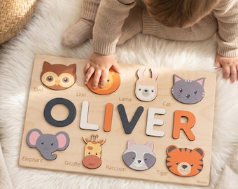 May include: A rectangular wooden name puzzle featuring the name OLIVER in large, multi-colored block letters. The board includes cutouts for animal faces, including an owl, lion, llama, cat, elephant, giraffe, raccoon, and tiger, with labels.