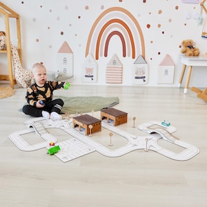 May include: A child plays with a wooden toy road system on a light wood floor. The road system includes a bridge, intersections, and two garages. The child is holding a small green toy car. The background features a rainbow wall decal and a playroom with a wooden rocking horse.
