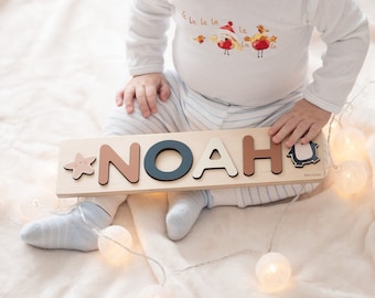 Puede incluir: Un rompecabezas de nombre de madera con el nombre "NOAH" deletreado en letras de colores. El rompecabezas está sobre una manta blanca con un bebé sentado a su lado.