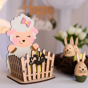 Puede incluir: Porta huevos de Pascua de madera con un lindo cordero con un lazo rosa. El soporte tiene un diseño de valla y dice "Feliz Pascua". También hay tres huevos de conejo de madera en un nido.