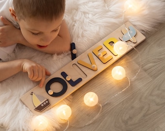 Puede incluir: Un niño juega con un rompecabezas de nombre de madera que deletrea "OLIVER". El rompecabezas tiene letras coloridas e ilustraciones de un velero y un avión. El rompecabezas está sobre una alfombra blanca y esponjosa con una cadena de luces blancas.