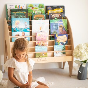 K&ouml;nnte beinhalten: Ein helles Holzregal mit mehreren horizontalen Ablagen, die verschiedene Kinderb&uuml;cher enthalten. Das Regal zeichnet sich durch ein geschwungenes Seitendesign aus und der Name Charlie ist in Schreibschrift auf der unteren Frontplatte eingraviert.
