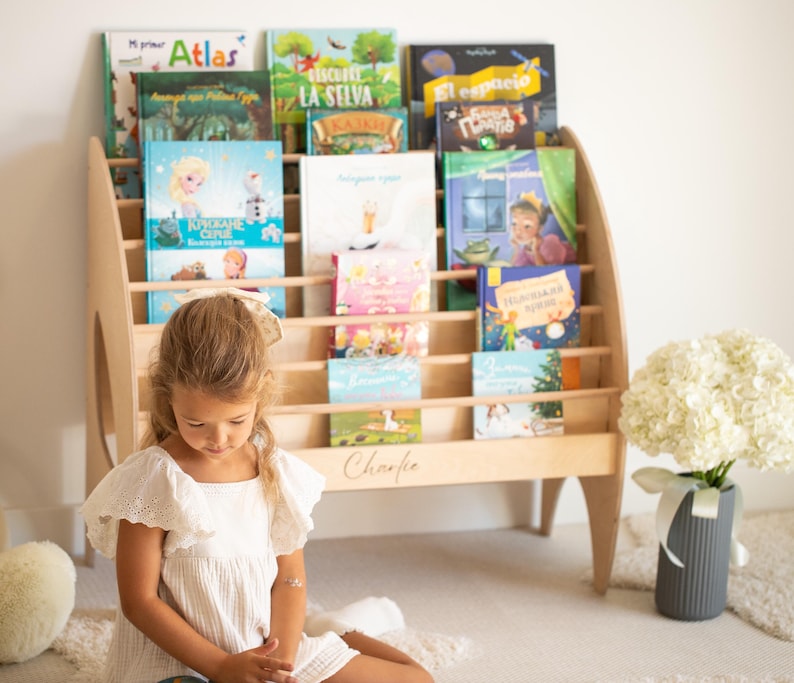 K&ouml;nnte beinhalten: Ein helles Holzregal mit mehreren horizontalen Ablagen, die verschiedene Kinderb&uuml;cher enthalten. Das Regal zeichnet sich durch ein geschwungenes Seitendesign aus und der Name Charlie ist in Schreibschrift auf der unteren Frontplatte eingraviert.