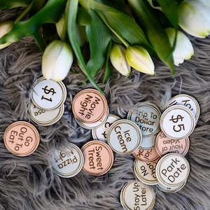 May include: A collection of round wooden tokens with various text and dollar amounts. The tokens are in shades of pink, blue, and white with brown lettering. The tokens are scattered on a grey fuzzy surface.