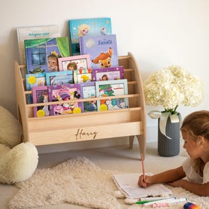 K&ouml;nnte beinhalten: Ein helles Bodenregal aus Holz mit horizontalen Leisten und dem eingravierten Namen Harry auf der Vorderseite. Das Regal ist mit verschiedenen Kinderb&uuml;chern gef&uuml;llt, die bunte Illustrationen und Figuren auf ihren Vorderseiten zeigen.