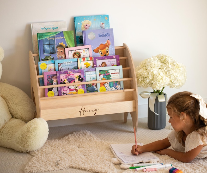 K&ouml;nnte beinhalten: Ein helles Bodenregal aus Holz mit horizontalen Leisten und dem eingravierten Namen Harry auf der Vorderseite. Das Regal ist mit verschiedenen Kinderb&uuml;chern gef&uuml;llt, die bunte Illustrationen und Figuren auf ihren Vorderseiten zeigen.