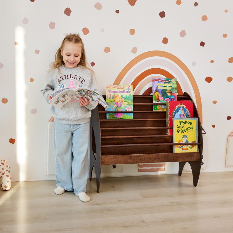 K&ouml;nnte beinhalten: Ein dunkelbraunes Bodenb&uuml;cherregal aus Holz mit vier horizontalen Ablagen, in denen Kinderb&uuml;cher mit den Titeln Thumbelina, Sleeping Beauty und Three Little Kittens stehen. Das Regal hat geschwungene Seitenteile und ein niedriges Design.