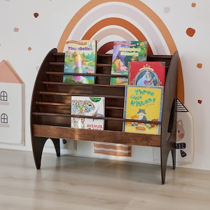 K&ouml;nnte beinhalten: Ein dunkelbraunes Holzregal mit vier horizontalen Ablagen zur Pr&auml;sentation von B&uuml;chern. Die Regale enthalten Kinderb&uuml;cher mit den Titeln Thumbelina, Sleeping Beauty, Animal Friends und Three Little Kittens. Das M&ouml;belst&uuml;ck hat geschwungene Seitenteile und Beine.