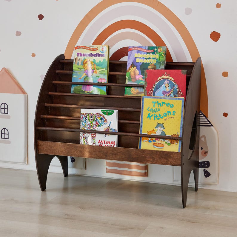 K&ouml;nnte beinhalten: Ein dunkelbraunes Holzregal mit vier horizontalen Ablagen zur Pr&auml;sentation von B&uuml;chern. Die Regale enthalten Kinderb&uuml;cher mit den Titeln Thumbelina, Sleeping Beauty, Animal Friends und Three Little Kittens. Das M&ouml;belst&uuml;ck hat geschwungene Seitenteile und Beine.