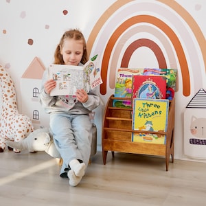K&ouml;nnte beinhalten: Ein h&ouml;lzernes Bodenregal mit drei horizontalen Ebenen, das mehrere Kinderb&uuml;cher enth&auml;lt, darunter ein Buch mit dem Titel Three Little Kittens, neben einem grauen elefantenf&ouml;rmigen Fu&szlig;hocker und einer Wand mit einem Regenbogen-Aufkleber.
