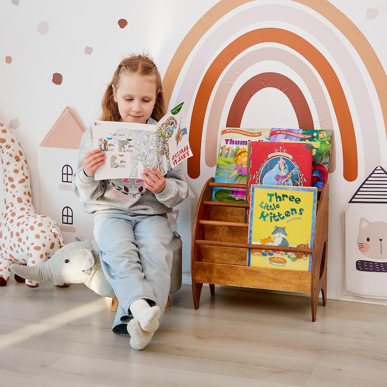 K&ouml;nnte beinhalten: Ein h&ouml;lzernes Bodenregal mit drei horizontalen Ebenen, das mehrere Kinderb&uuml;cher enth&auml;lt, darunter ein Buch mit dem Titel Three Little Kittens, neben einem grauen elefantenf&ouml;rmigen Fu&szlig;hocker und einer Wand mit einem Regenbogen-Aufkleber.