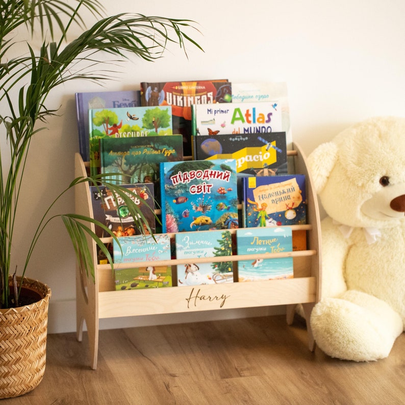 K&ouml;nnte beinhalten: Ein helles Holzregal mit drei horizontalen Ebenen zur Pr&auml;sentation von B&uuml;chern. Auf der Vorderseite ist der Name Harry in Schreibschrift eingraviert. Die Regale sind mit verschiedenen Kinderb&uuml;chern mit bunten, illustrierten Einb&auml;nden gef&uuml;llt.