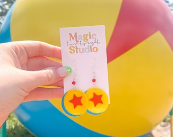 Könnte beinhalten: Ein Paar gelbe und rote Ohrringe mit einem Sternendesign. Die Ohrringe befinden sich vor einem großen gelben, roten und blauen Ball. Der Text "Magic twenty-eight Studio" steht auf einer weißen Karte.
