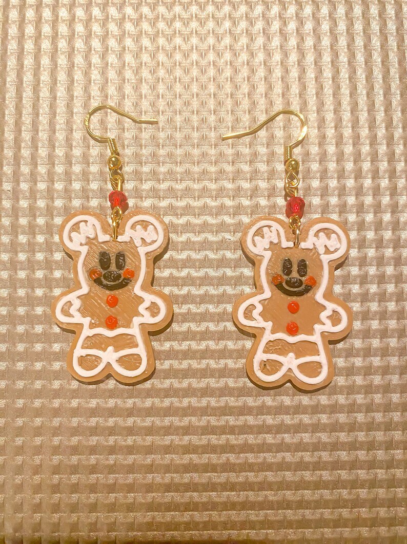 Op de afbeelding: Een paar gouden oorhangers met gingerbread man-bedels met witte glazuurdetails en rode knopen. De gingerbread men hebben zwarte ogen en een glimlachende mond.