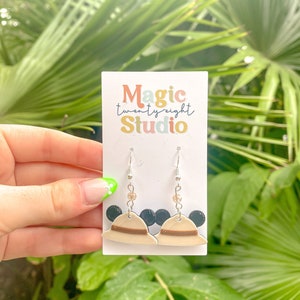 May include: A pair of dangle earrings featuring a brown and beige hat with black Mickey Mouse ears. The earrings are on a white card with the text "Magic twenty eight Studio".