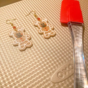 Op de afbeelding: Een paar gingerbread man oorbellen met zwarte ogen en rode accenten op een gouden bakplaat. De oorbellen hangen aan gouden haken.