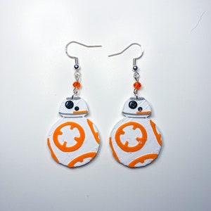 May include: Pair of white and orange BB-8 droid earrings. The earrings feature a round design with orange circular patterns and a silver hook. Each earring has a small orange bead accent.