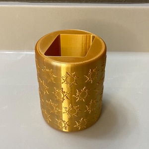 May include: A cylindrical, gold-colored container with a square opening and sunburst pattern. The container is likely 3D-printed and sits on a white surface. The sunburst design is repeated around the exterior of the container.