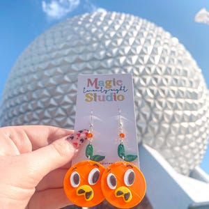 May include: Orange character earrings with a green leaf, inspired by a Disney character. The earrings are on a card that says "Magic twenty eight Studio".
