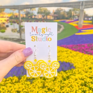 May include: A pair of yellow lemon-shaped earrings with white centers, hanging from silver hooks. The earrings are in the shape of Mickey Mouse ears. The earrings are in front of a card that says "Magic twenty eight Studio".
