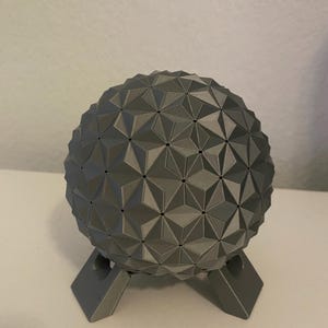 May include: A gray, geometric sphere with a complex, textured surface, resting on a matching gray stand. The sphere is composed of numerous triangular facets, creating a unique, architectural design. The stand has a simple, angular design.