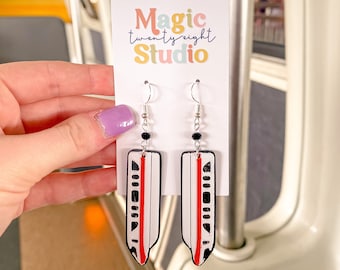 May include: A pair of white, red, and black dangle earrings featuring a stylized image of a monorail train. The earrings are hanging from a card that reads "Magic twenty-eight Studio".