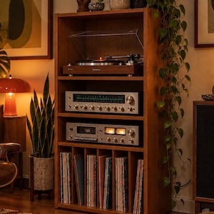 May include: A tall wooden media storage unit with four shelves. The top shelf holds a Technics turntable, the middle shelves house a silver stereo receiver and a cassette deck, and the bottom shelf features vertical dividers for vinyl records.