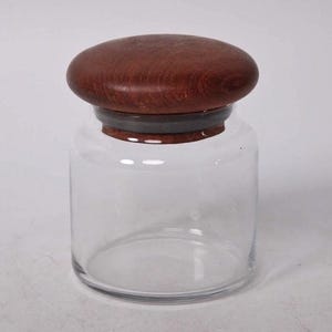 Puede incluir: Un tarro de cristal transparente con tapa de madera marrón oscuro. La tapa es redonda y plana, con veta de madera visible. El tarro tiene forma cilíndrica y un anillo gris oscuro entre el cristal y la tapa. El tarro está vacío.