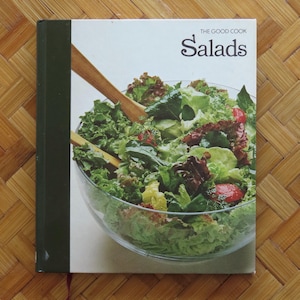 Op de afbeelding: Een kookboek getiteld "The Good Cook Salads" met een groene rug en een witte kaft. De kaft toont een grote glazen kom gevuld met een kleurrijke salade, houten saladebestek en de titel in zwarte letters.