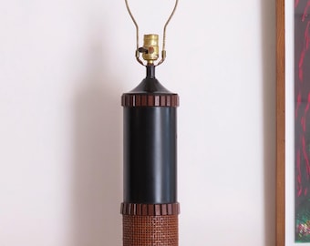 May include: A black and brown cylindrical table lamp with a woven rattan base and accents. The lamp has a brass-coloured neck and a metal shade holder. The lamp is approximately 61 cm tall.