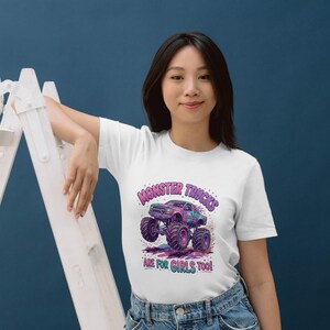 May include: White t-shirt featuring a colorful graphic of a monster truck with the text "MONSTER TRUCKS ARE FOR GIRLS TOO!". The design includes shades of purple, blue, and green. The shirt is made of a soft material.