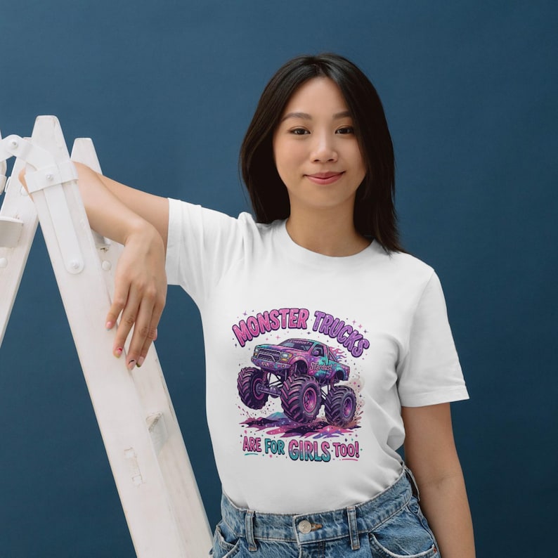 May include: White t-shirt featuring a colorful graphic of a monster truck with the text "MONSTER TRUCKS ARE FOR GIRLS TOO!". The design includes shades of purple, blue, and green. The shirt is made of a soft material.