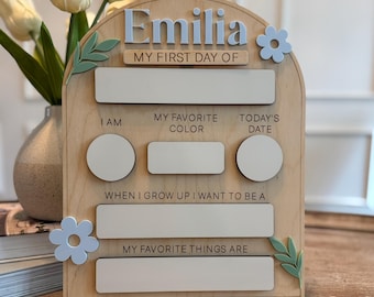 May include: A wooden first day of school sign, featuring the name "Emilia" in light blue. The sign includes spaces for "I am", "My favourite colour", "Today's date", "When I grow up I want to be a", and "My favourite things are". Decorative flowers and leaves are also present.