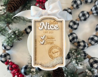Puede incluir: Un adorno navideño de madera con forma de lista con las palabras "Santa's Nice List 2023" y dos nombres marcados: Jaxson y Joseph. El adorno tiene una cinta blanca y un sello de North Pole Workshop.